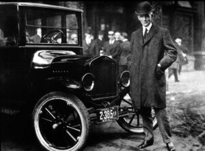 photo of Henry Ford with Model T, Hotel Iroquois, Buffalo NY 1921, this work is in the public domain in the United States because it was published (or registered with the U.S. Copyright Office) before January 1, 1931.